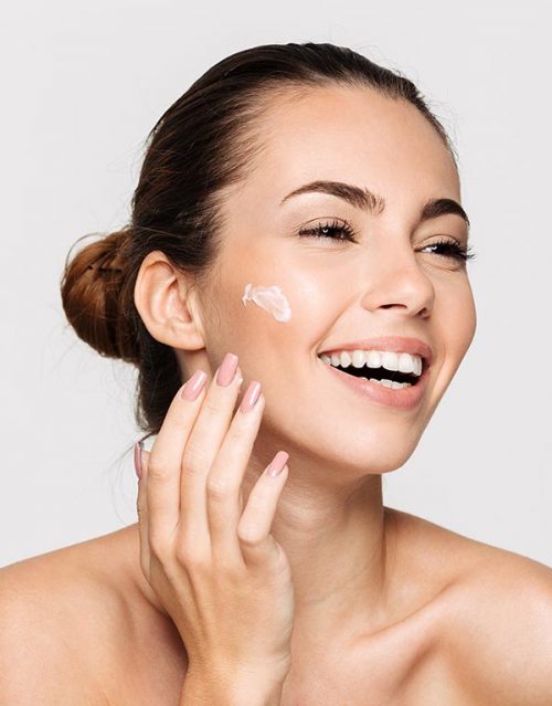 Close up beauty portrait of a laughing beautiful half naked woman applying face cream and looking away isolated over white background